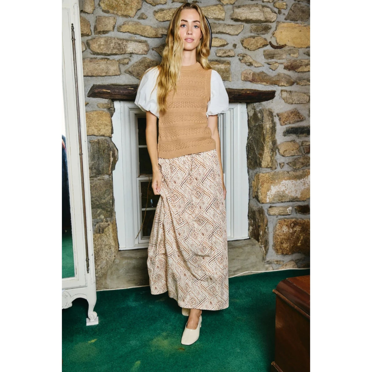 A woman stands indoors in front of a stone wall and window, wearing a tan sleeveless knit top over a white short-sleeve blouse, a beige patterned maxi skirt, and white pointed shoes.