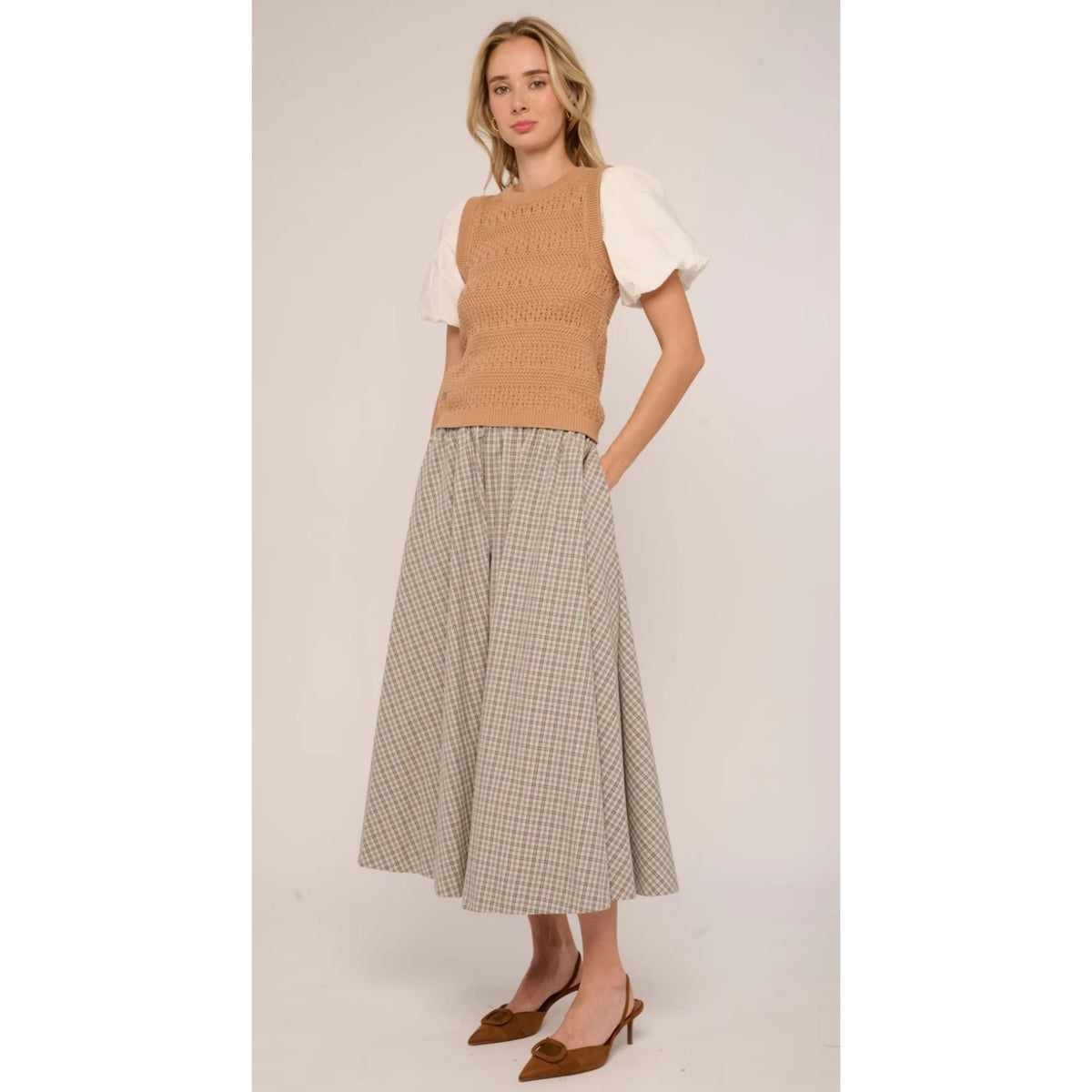 A woman modeling a sleeveless tan knit vest over a white puff-sleeve top, paired with a beige and brown checkered midi skirt and brown pointed-toe flats, against a plain light beige studio background.
