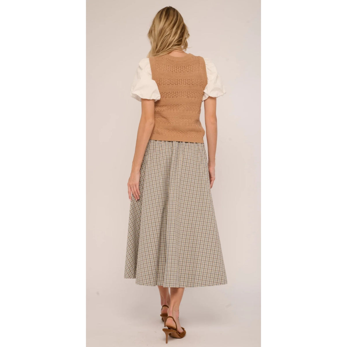 A woman shown from the back wearing a tan knit sleeveless vest over a white puff-sleeve top and a beige-and-gray checkered midi skirt, paired with brown heels, against a plain light beige studio background.