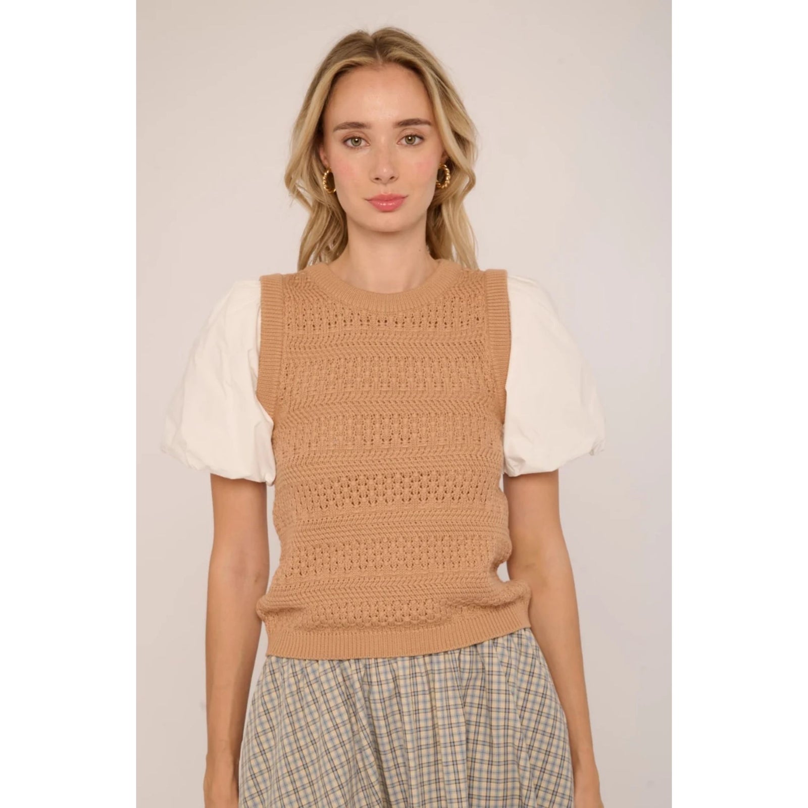 A woman modeling a sleeveless tan knit sweater vest layered over a white puff-sleeve top and beige plaid trousers, set against a plain light beige studio background.