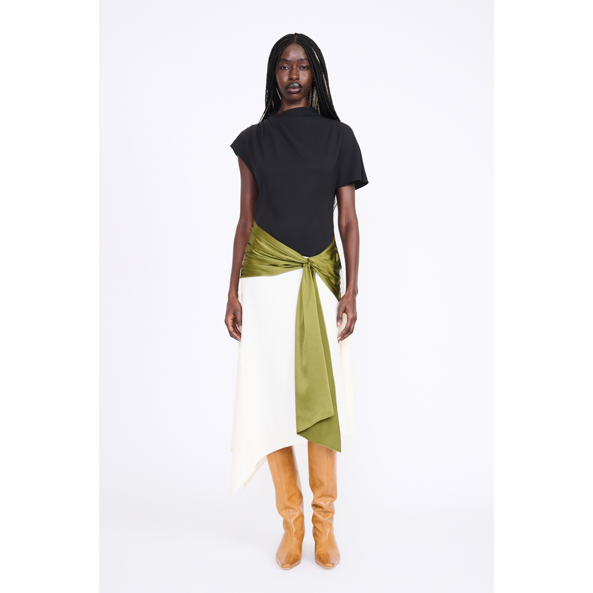 A model is shown from the back wearing a black short-sleeve top and a white asymmetrical skirt with an olive green satin sash tied at the waist, paired with tan boots, against a plain white studio background.