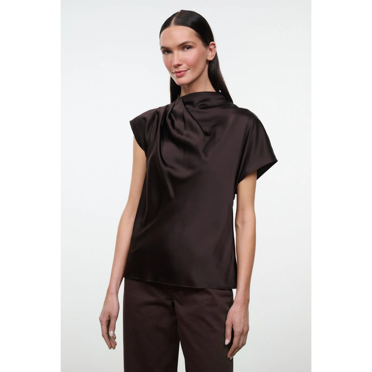 A woman modeling a dark brown satin-like short-sleeve top with a draped, gathered high neckline, paired with matching dark brown trousers, against a plain light gray studio background.