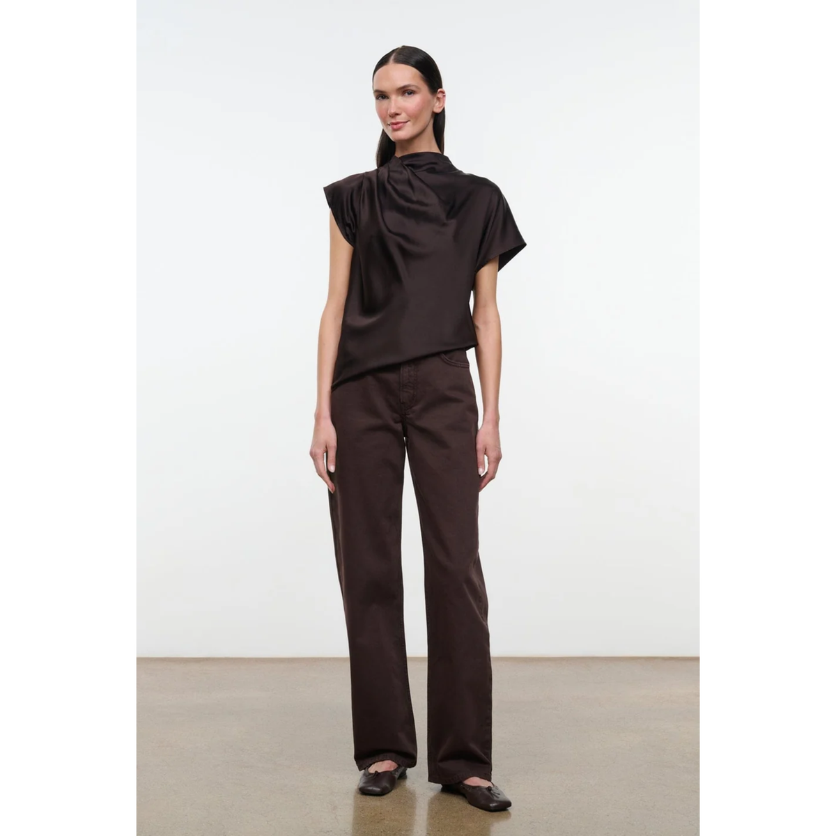 A model stands in a studio wearing a dark brown draped short-sleeve top and matching dark brown wide-leg trousers with black flats.