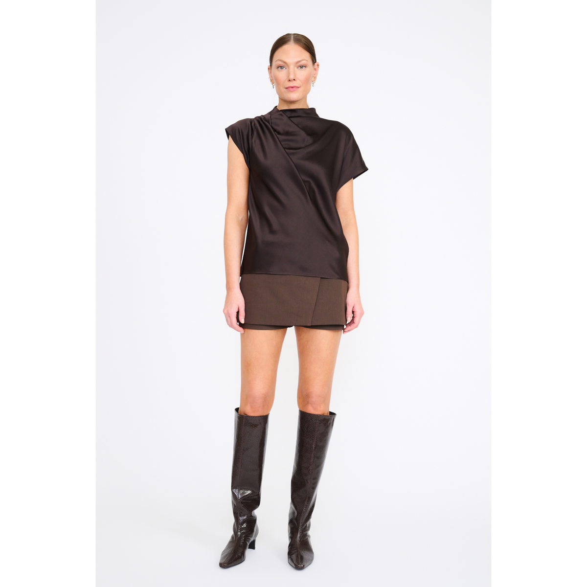 A model stands against a light gray studio background wearing a dark brown draped short-sleeve top and matching brown mini skirt, paired with tall black knee-high boots.