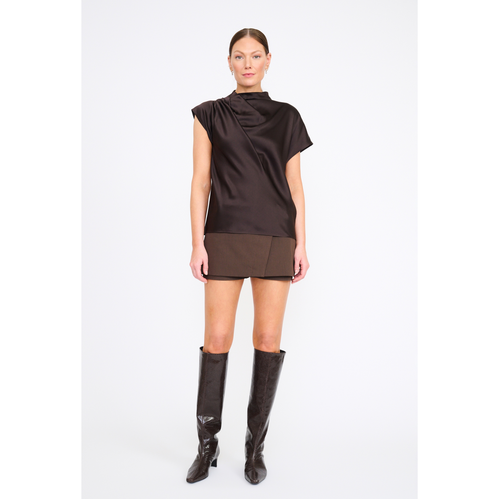 A model stands against a light gray studio background wearing a dark brown draped short-sleeve top and matching brown mini skirt, paired with tall black knee-high boots.