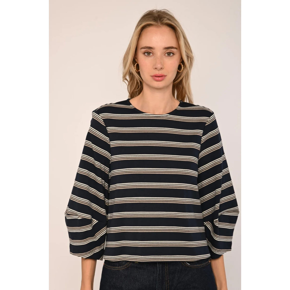 A woman wearing a navy blue and beige striped top with three-quarter sleeves, paired with dark jeans, set against a plain light beige background.