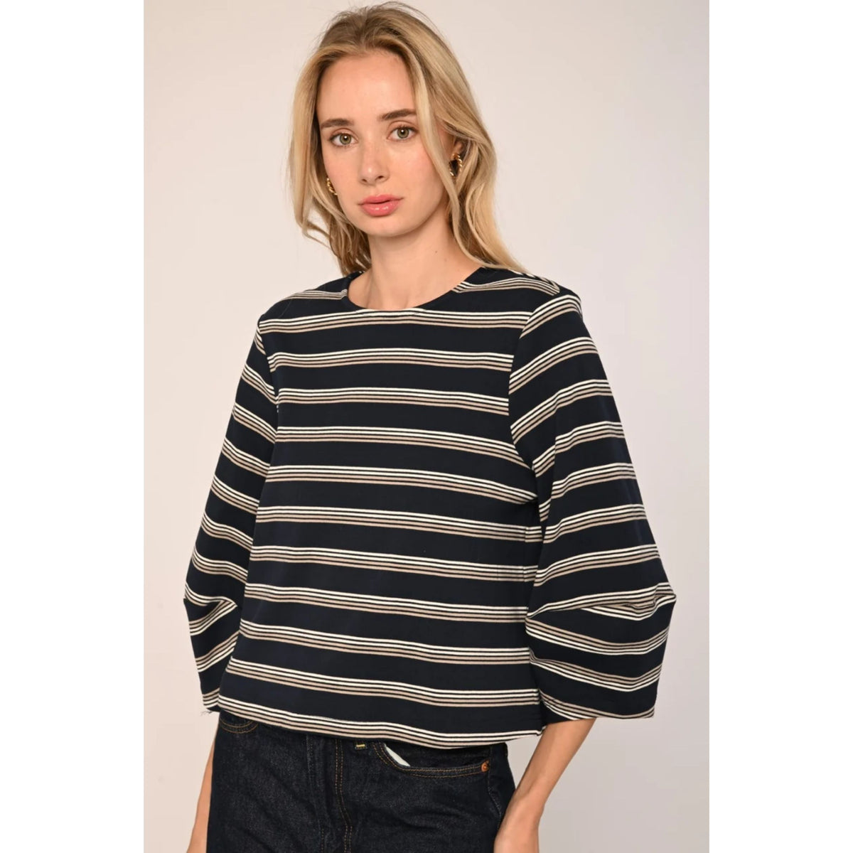 A woman wearing a loose-fitting, short-sleeved striped top with horizontal navy, beige, and white stripes, paired with dark jeans, set against a plain light beige studio background.