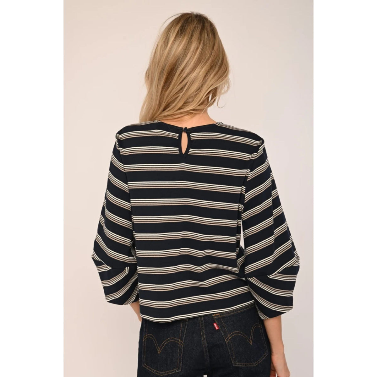 A woman shown from the back wearing a black and beige horizontally striped long-sleeve top with a small keyhole cutout at the neckline, paired with dark denim jeans.