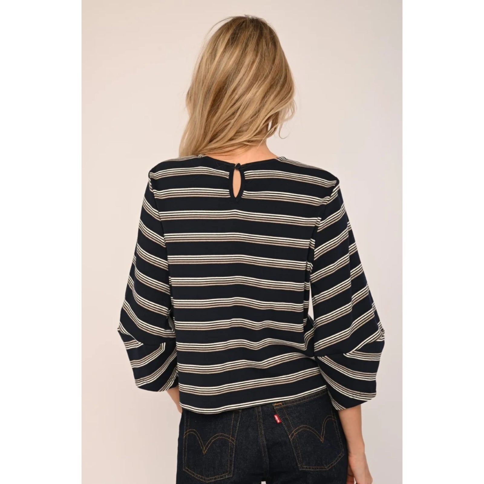 A woman wearing a navy blue and beige striped top with three-quarter sleeves, paired with dark jeans, set against a plain light beige background.