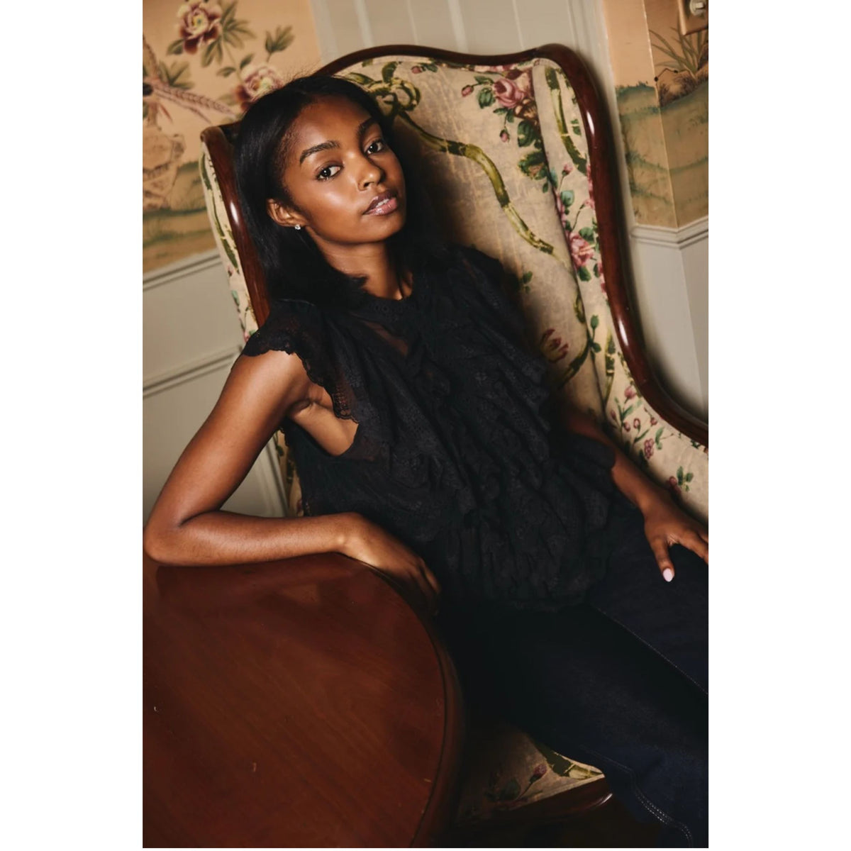 A woman wearing a black lace sleeveless top and dark pants reclines in a vintage floral upholstered armchair next to a wooden table in an indoor setting.