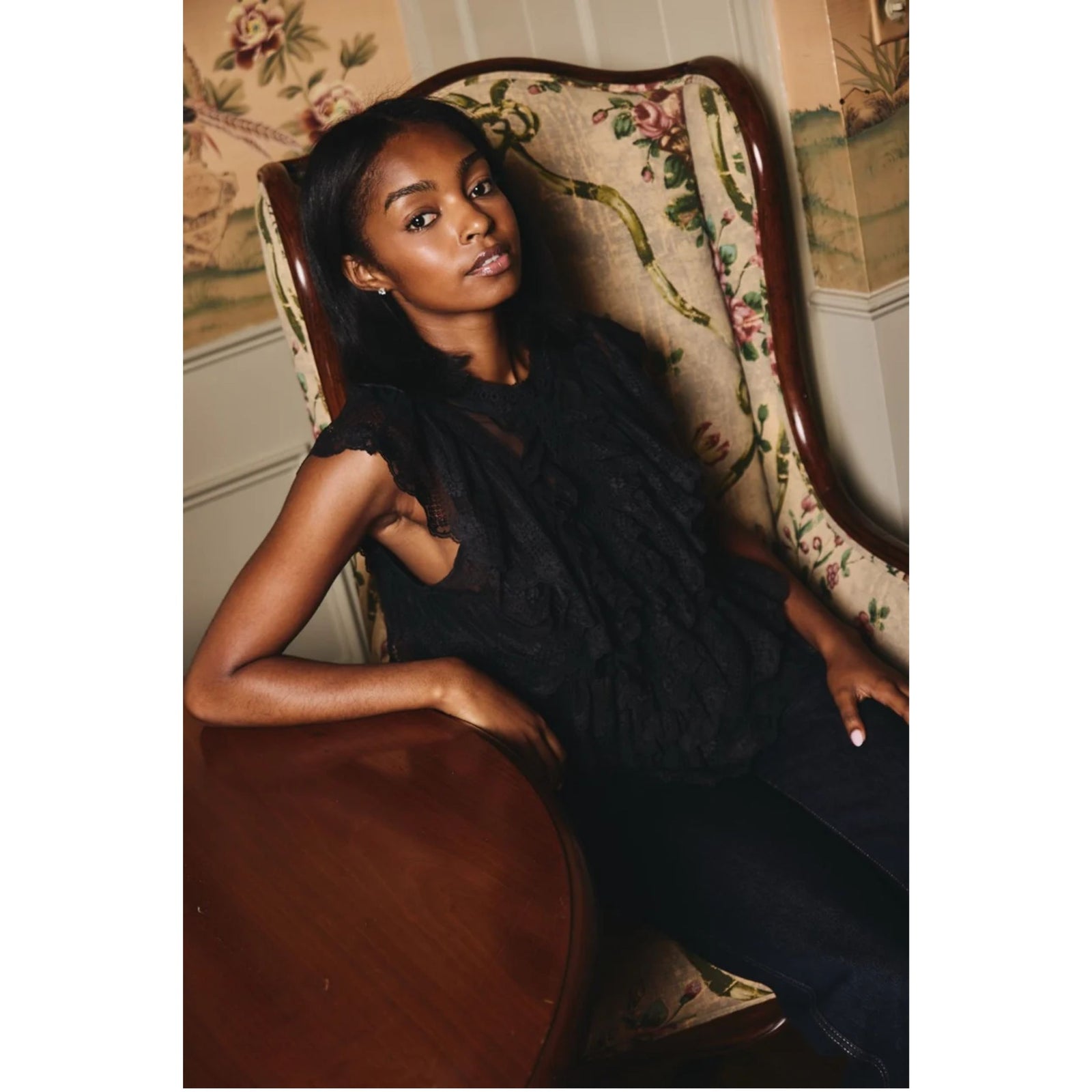 A woman wearing a black lace sleeveless top and dark pants reclines in a vintage floral upholstered armchair next to a wooden table in an indoor setting.