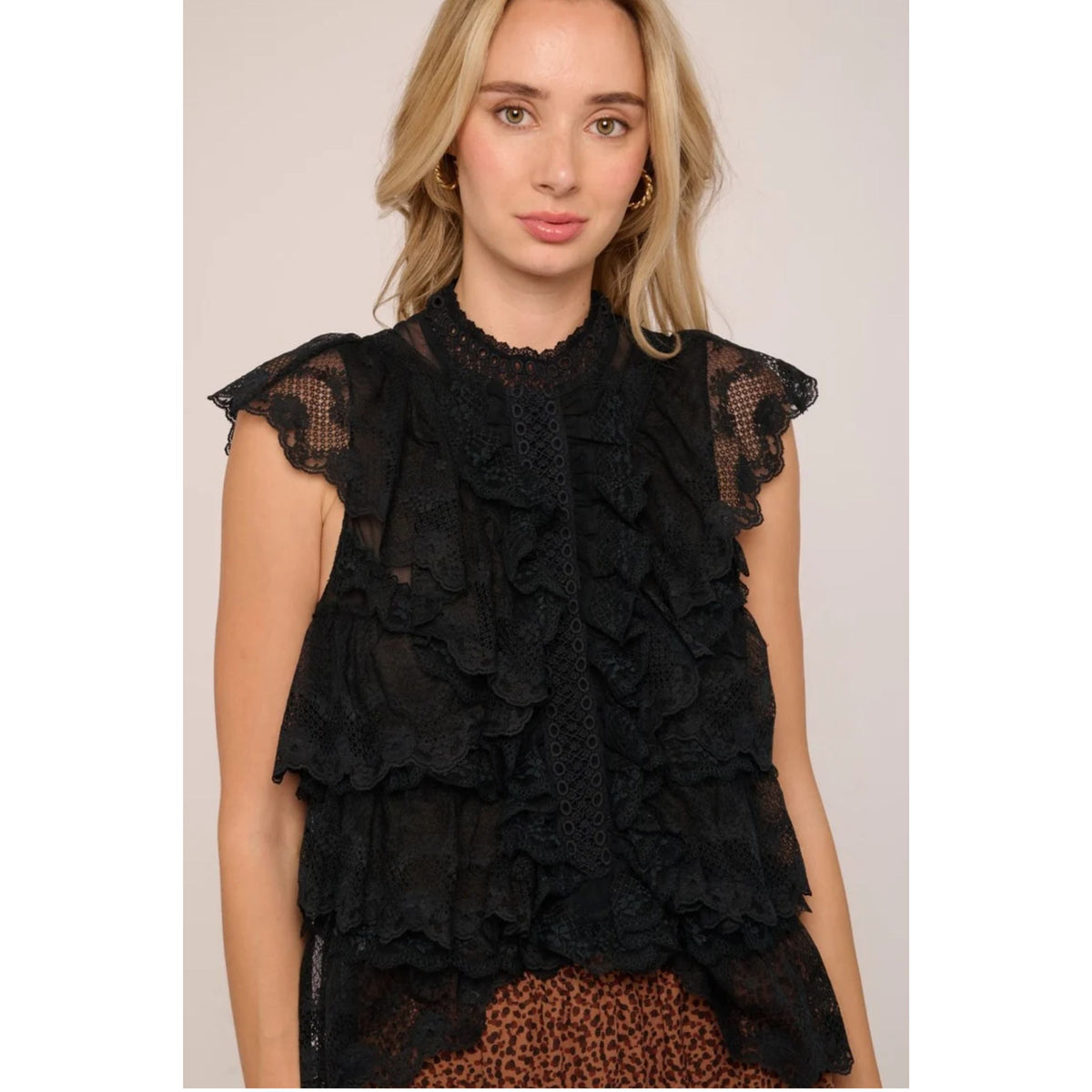 A woman wearing a black ruffled lace sleeveless top with sheer cap sleeves, paired with a brown leopard-print bottom, against a light beige studio background.