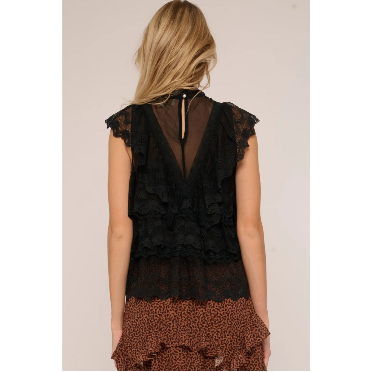 A woman shown from the back wearing a sleeveless black ruffled lace top with a sheer back panel and a brown leopard-print ruffled hem, against a plain light beige studio background.