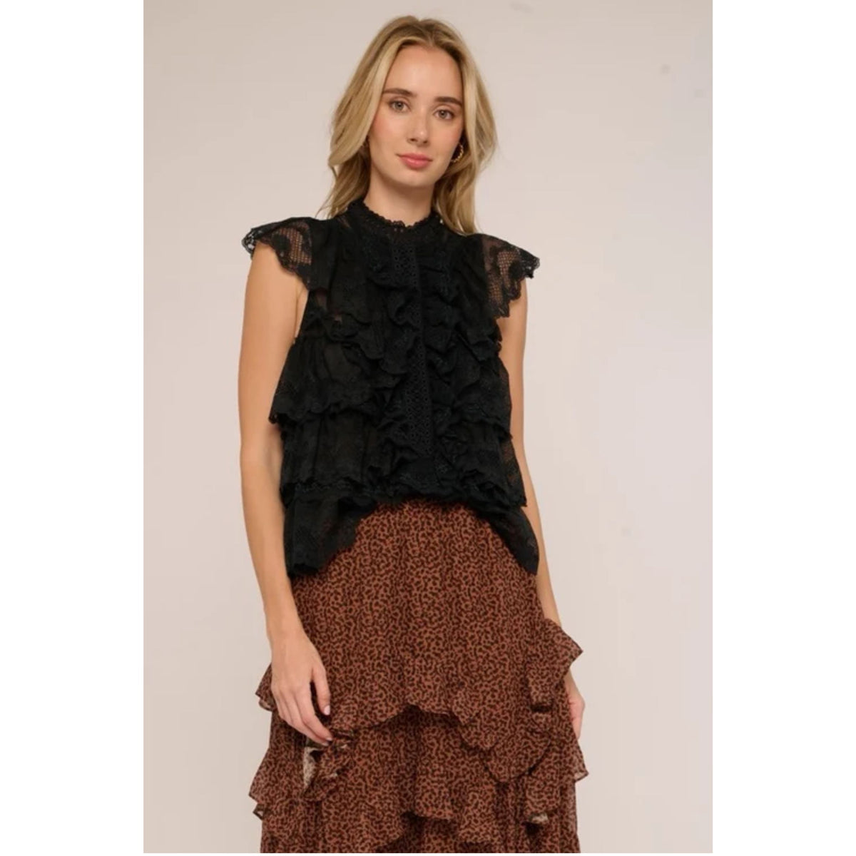 A woman modeling a black ruffled sleeveless top paired with a brown patterned ruffled skirt, photographed against a plain light beige studio background.