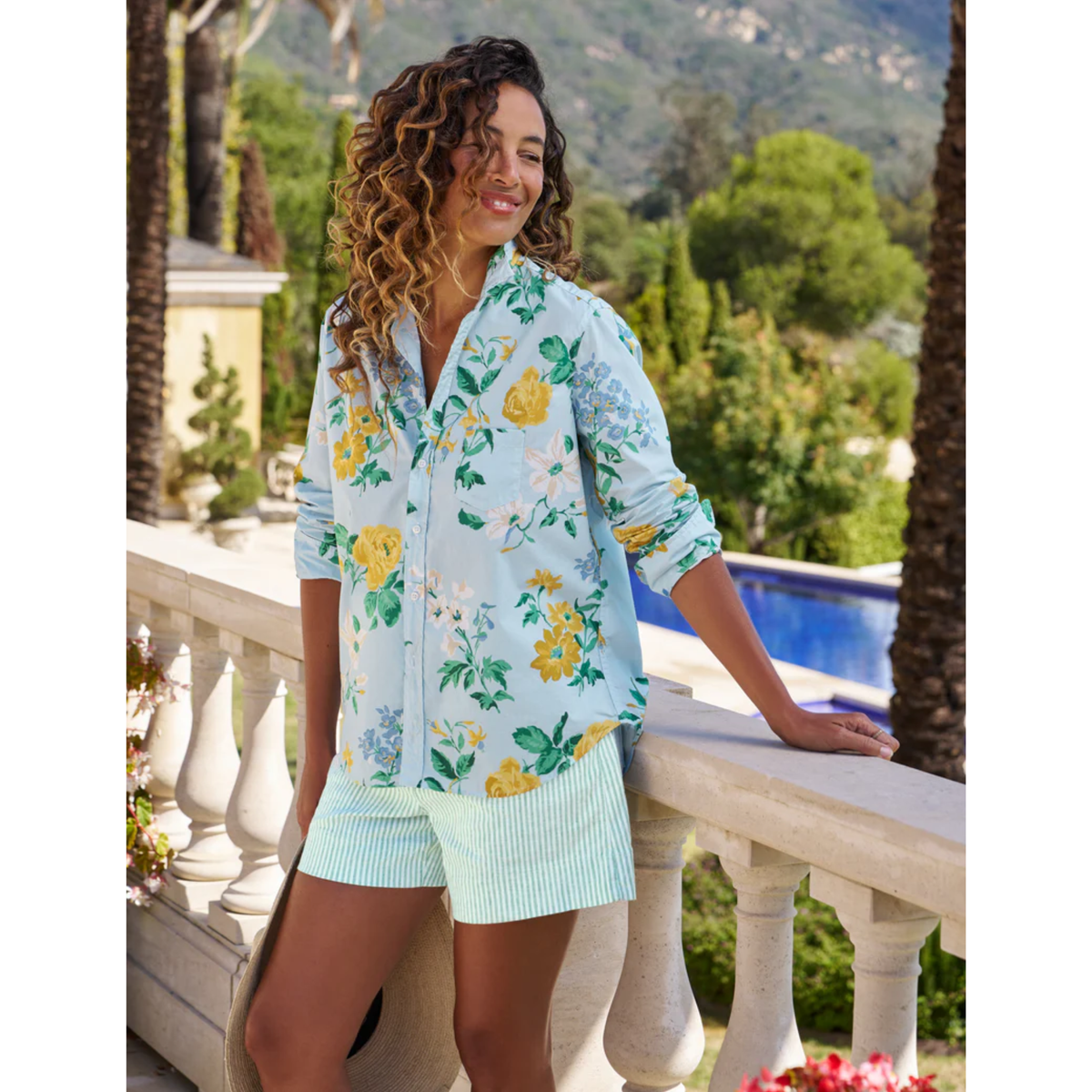 Woman wearing a floral shirt and shorts standing on a balcony with greenery and mountains in the background