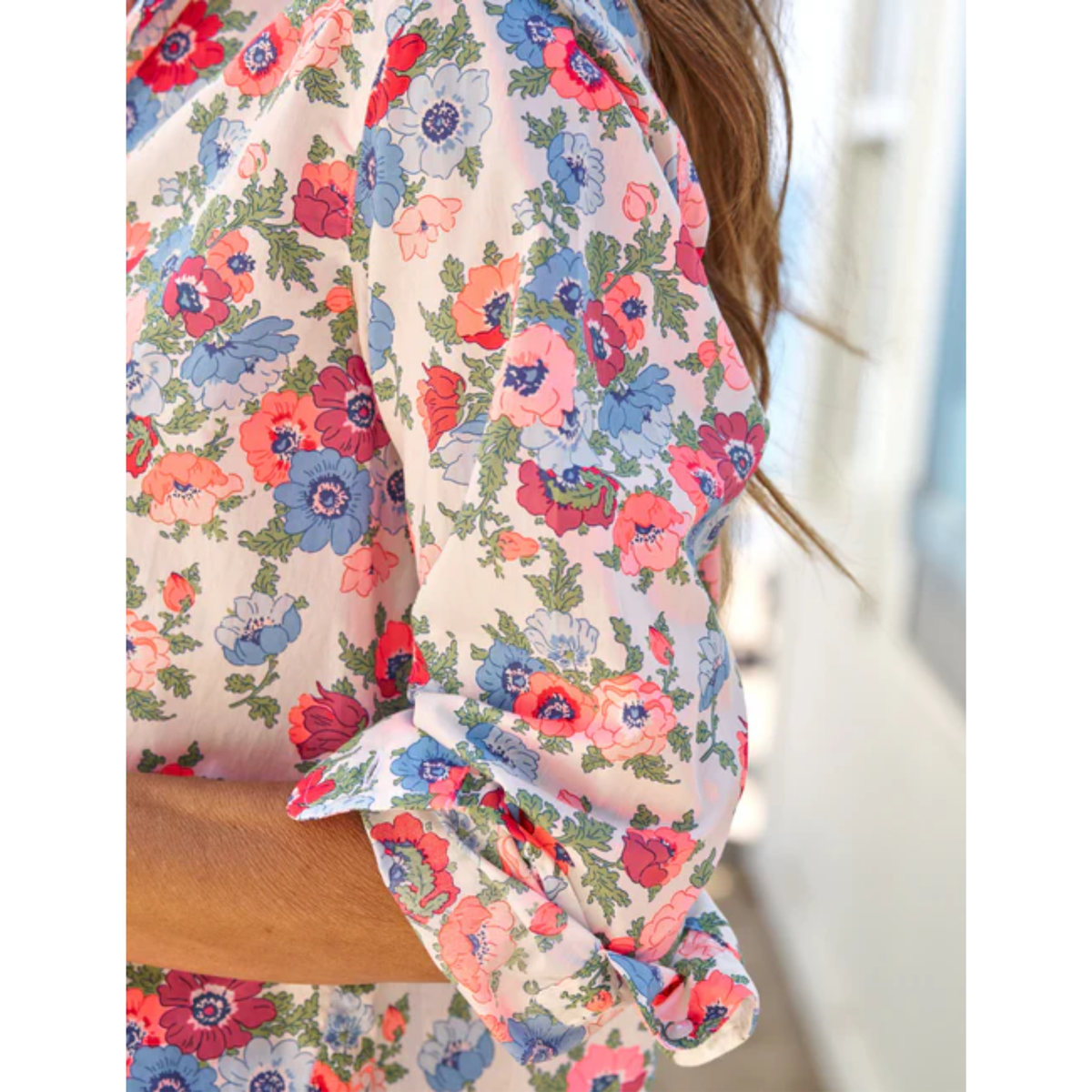Close-up of a woman wearing a long-sleeve blouse with a colorful floral print on a light cream background. The blouse features pink, blue, and red flowers with green leaves, and the sleeve is rolled up with a ruffled cuff.
