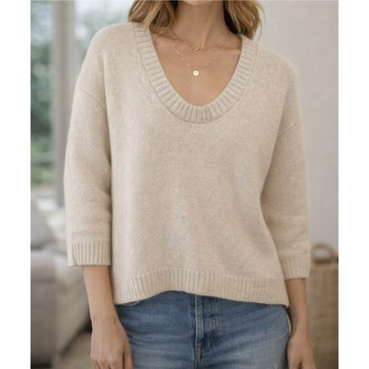 A woman wearing a light beige knit sweater with a wide ribbed v-neck and three-quarter sleeves, paired with blue jeans.