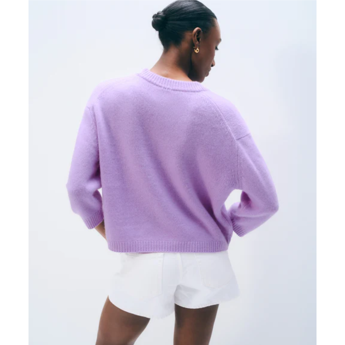 A woman shown from the back wearing a light purple knit sweater and white shorts against a plain light gray studio background.