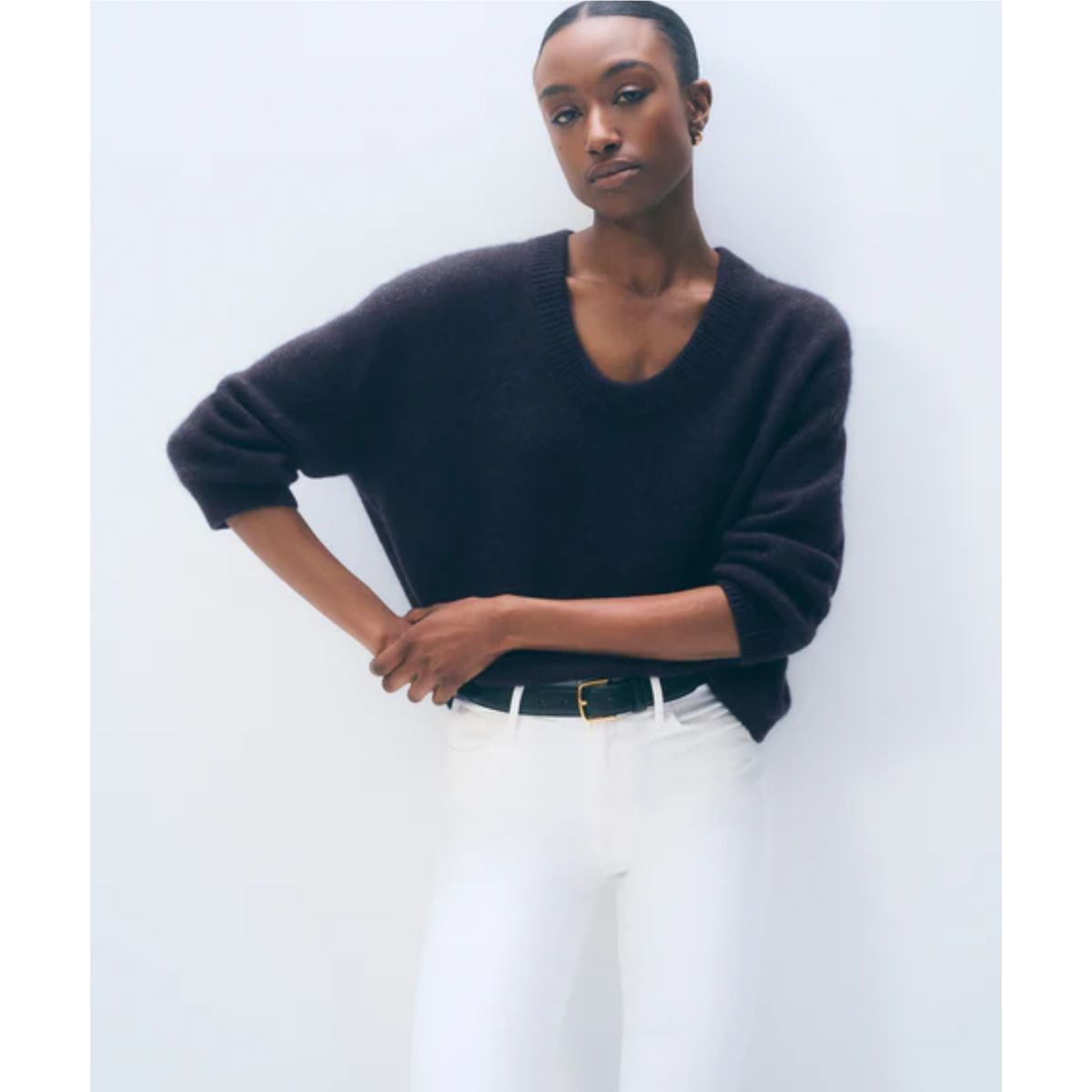 A model wearing a dark charcoal knit sweater, white trousers, and a black belt, standing against a light gray studio background.