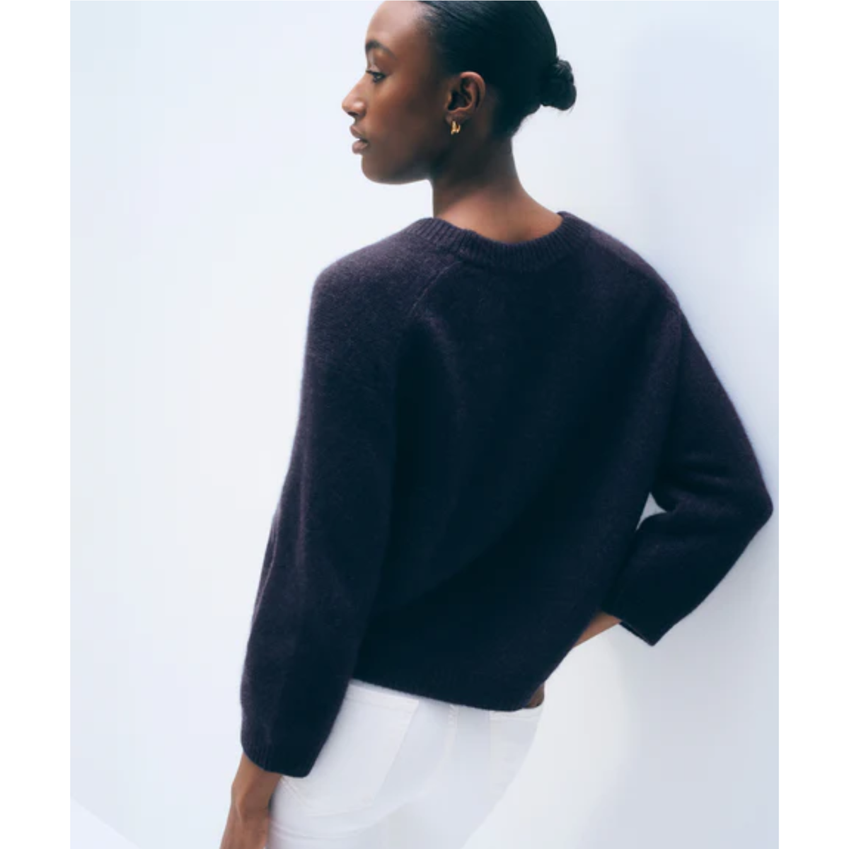 A woman shown from the back wearing a dark charcoal knit sweater and white pants, standing against a plain light gray studio background.