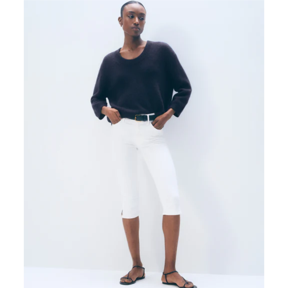 A model wearing a black long-sleeve knit sweater, white cropped pants, and black strappy sandals, standing against a plain white studio background.