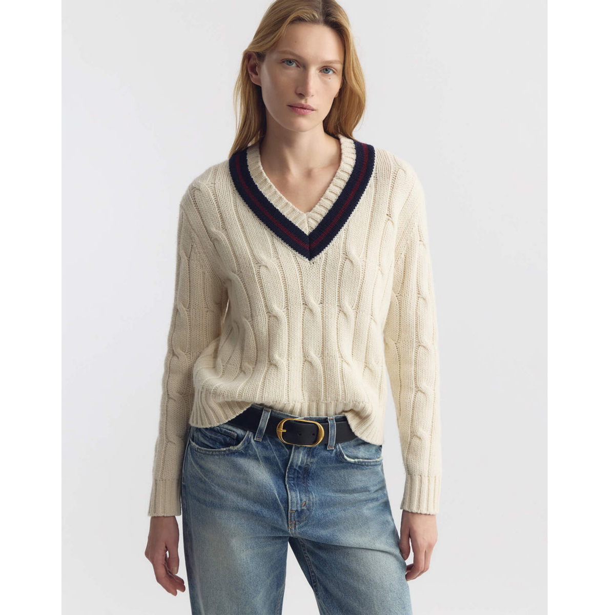 Woman wearing a cream cable knit sweater with a dark collar and blue jeans on a white background
