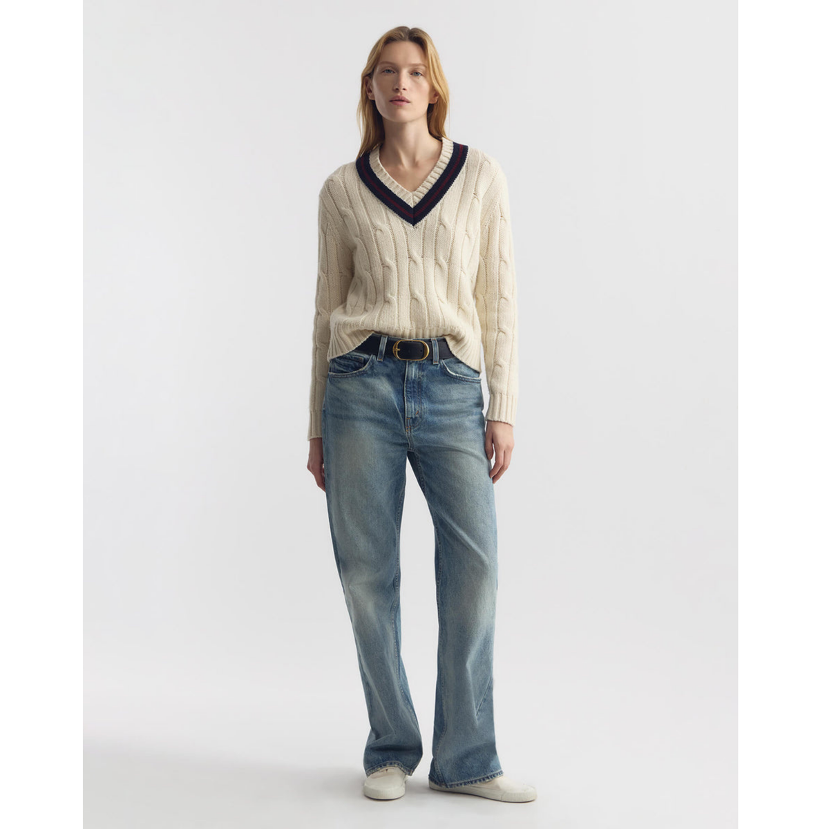 Woman wearing a cream sweater with a dark V-neckline and blue jeans on a white background