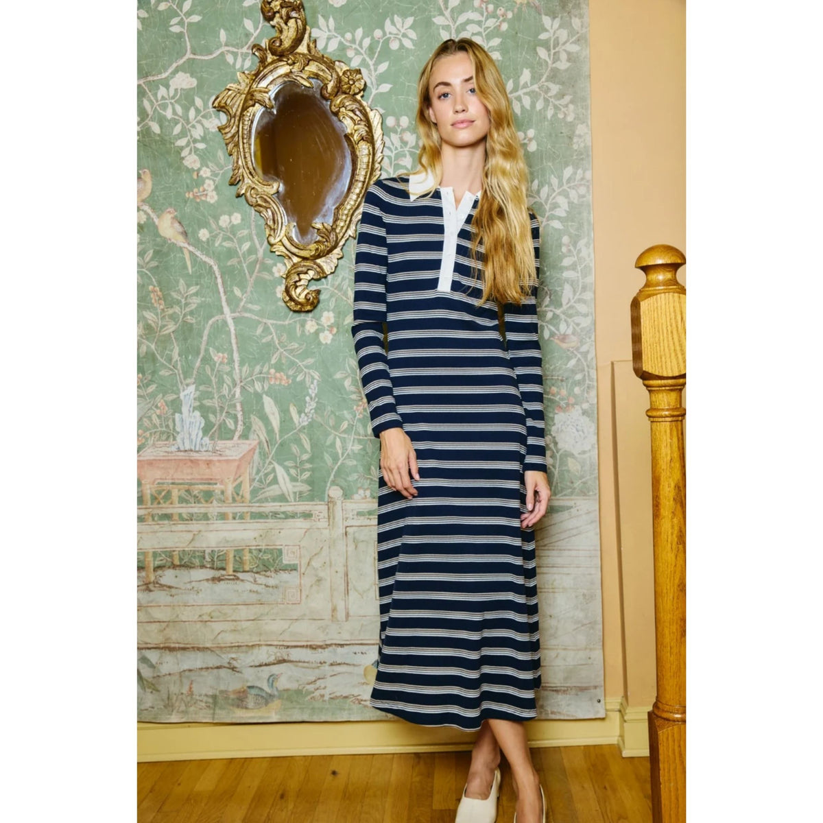 A woman modeling a navy and white horizontally striped long-sleeve midi dress with a white collar, standing in a room with vintage floral wallpaper, a gold mirror, and a wooden staircase railing.