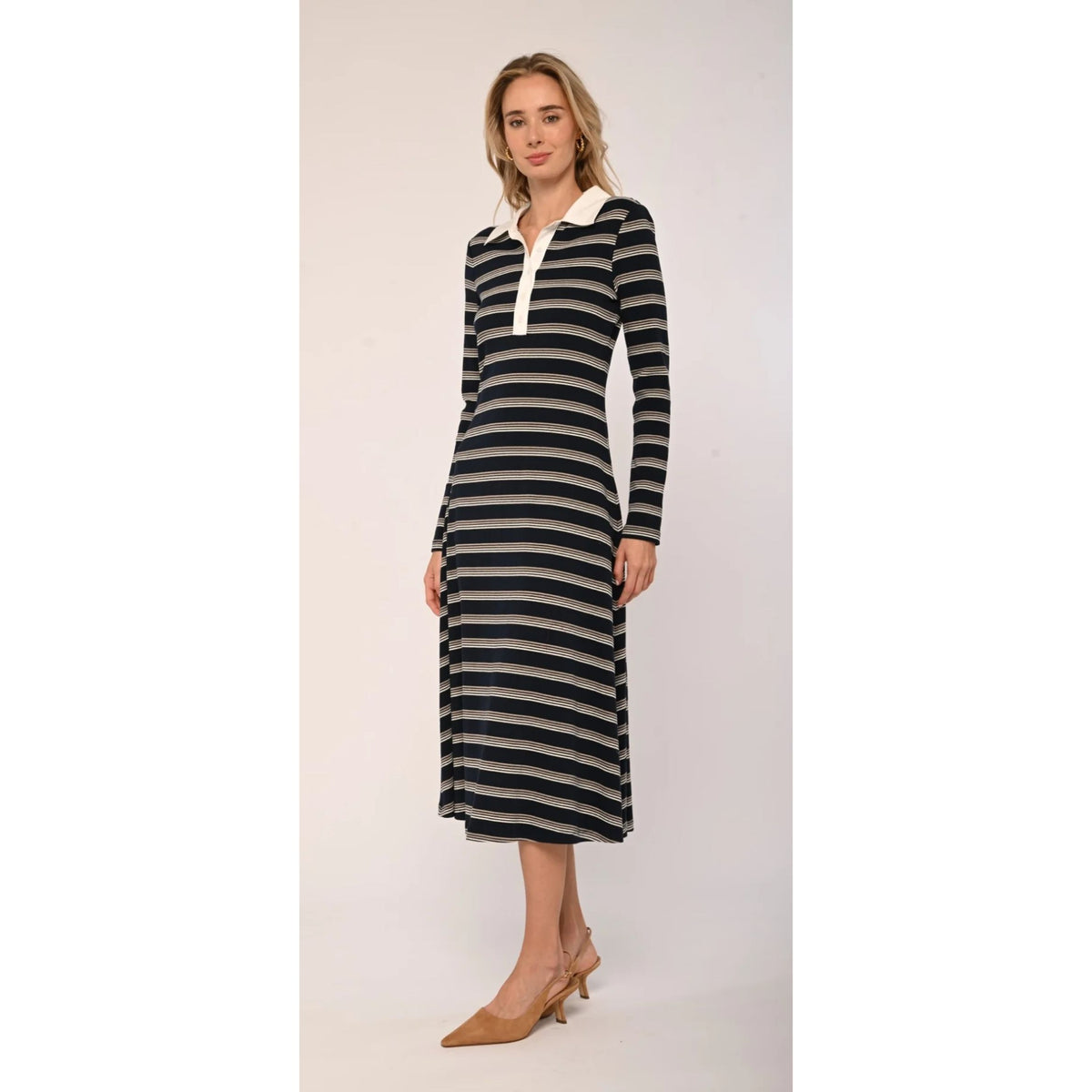 A woman modeling a long-sleeve midi dress with horizontal black and beige stripes, featuring a white collar and a white button placket, paired with beige pointed-toe heels against a plain light background.