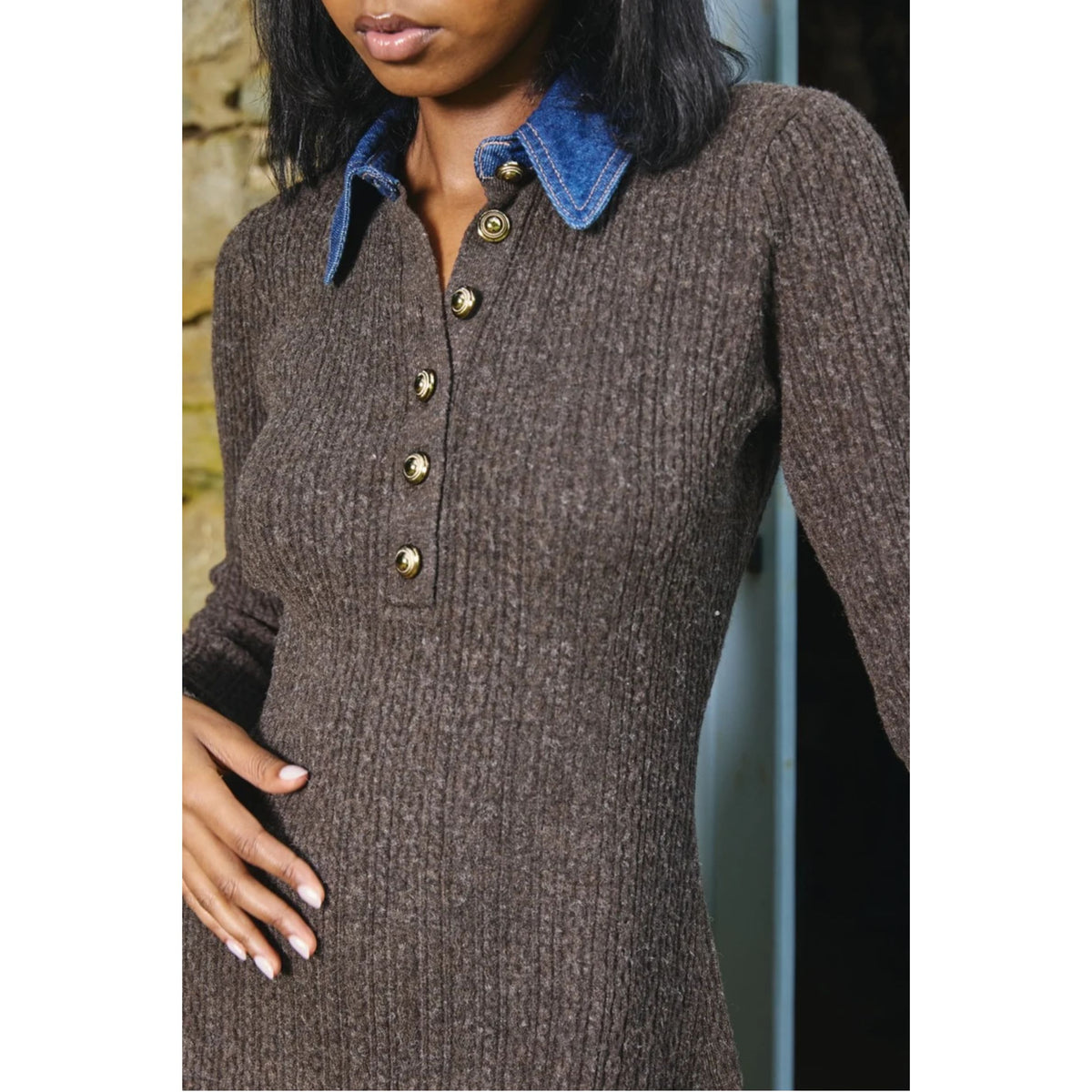 A woman wearing a brown ribbed knit long-sleeve sweater dress with a blue denim collar and gold buttons, shown from the lips down against a stone wall background.