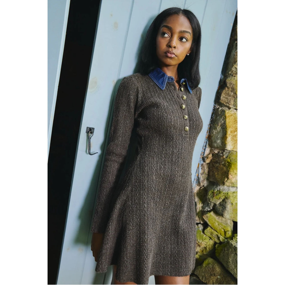 A woman wearing a brown ribbed knit long-sleeve dress with a blue denim-style collar and gold buttons, standing next to a light blue door and a stone wall.