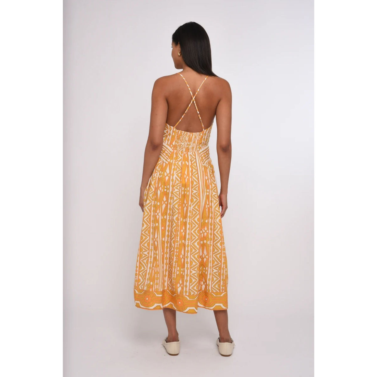 A woman viewed from behind wearing a yellow and white patterned sleeveless maxi dress with crisscross back straps, paired with beige flat shoes, against a light gray studio background.