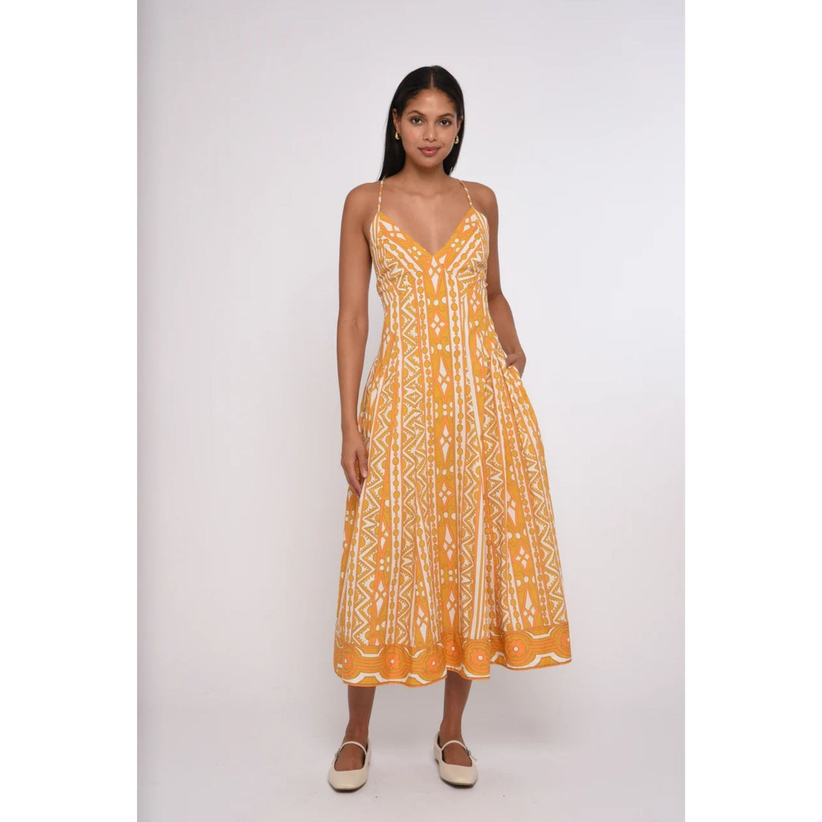 A woman modeling a sleeveless midi dress with thin straps and a white geometric tribal-style pattern on a bright yellow-orange base, paired with beige flat shoes, against a light gray studio background.
