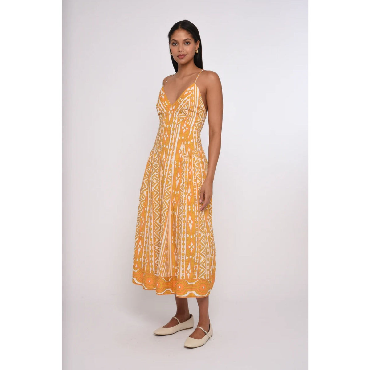A woman modeling a yellow and white patterned midi dress with thin straps and a V-neckline, paired with beige flat shoes, against a plain light gray studio background.