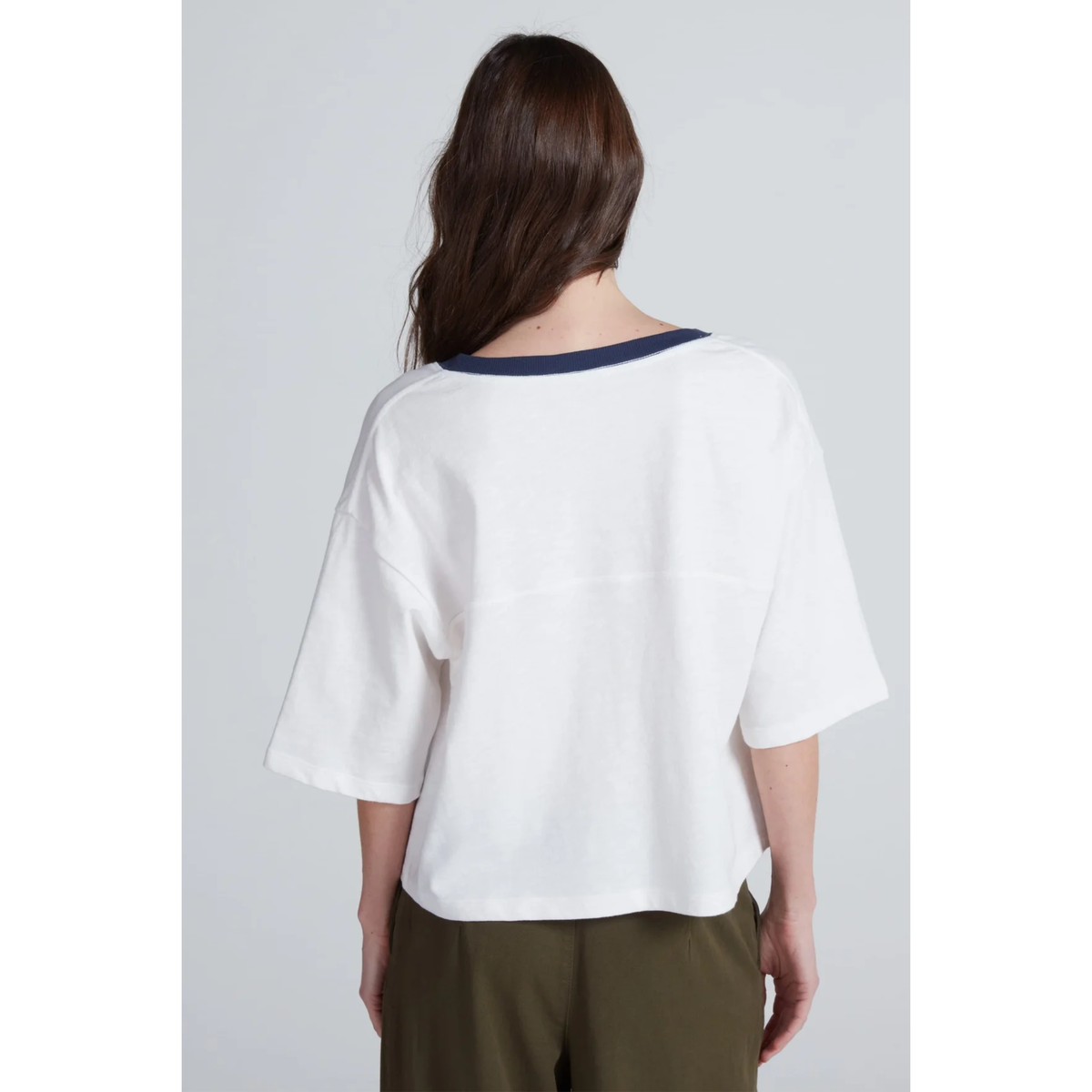 A woman viewed from the back wearing a white short-sleeve top with a dark navy blue collar trim, paired with olive green pants, against a light gray studio background.