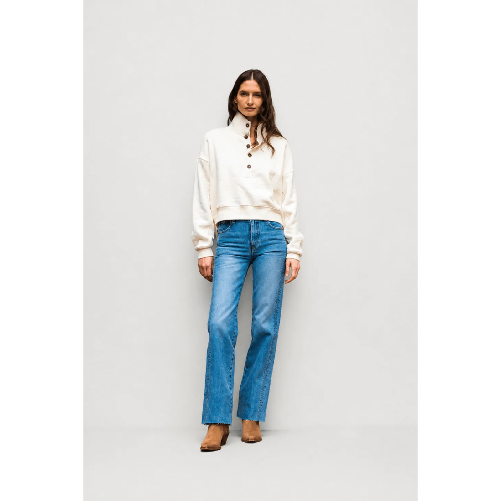 A woman stands against a light gray studio background wearing a cream cropped henley sweatshirt, blue straight-leg jeans, and brown boots.