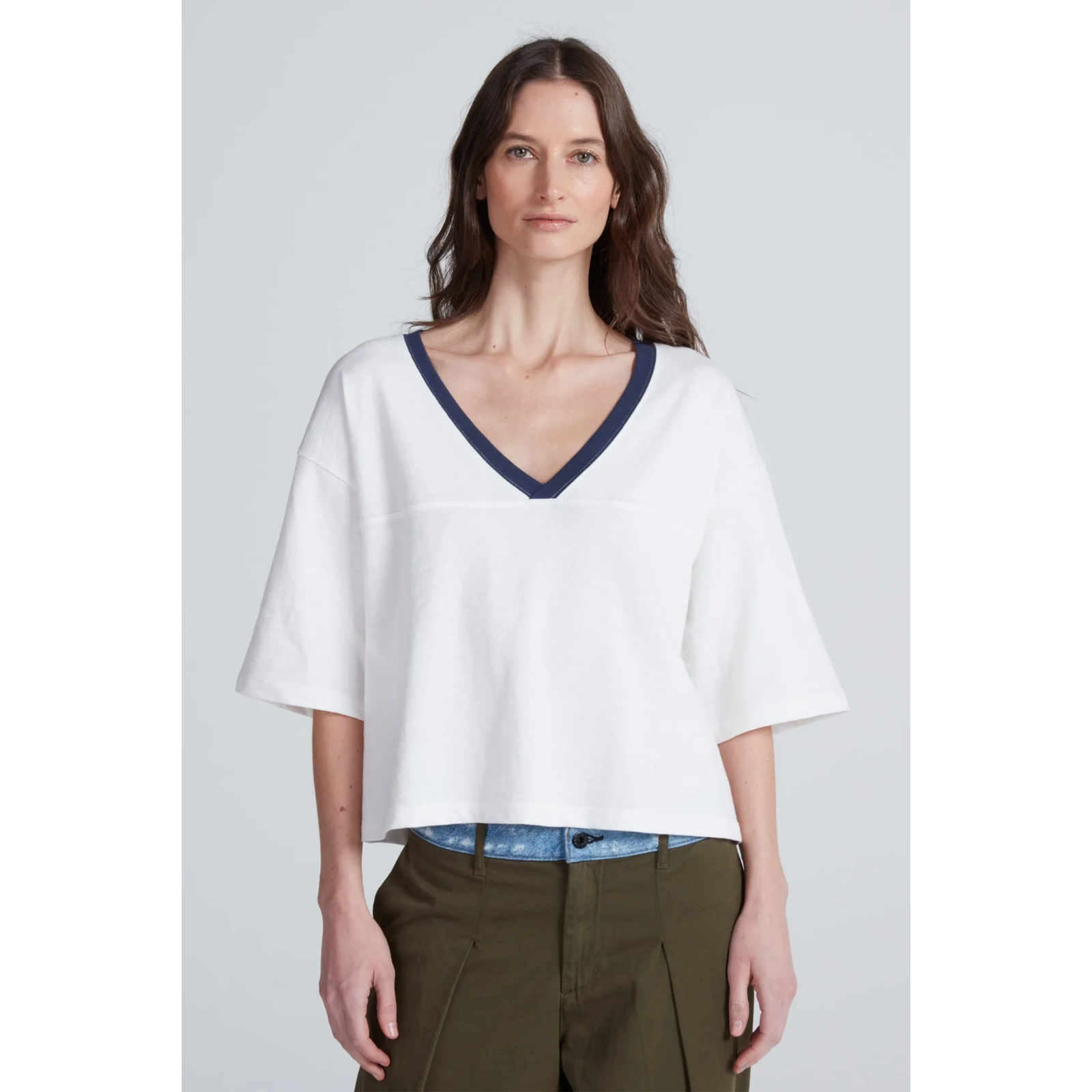 A woman modeling a white short-sleeve V-neck top with dark navy trim, paired with olive green pants, against a light gray studio background.