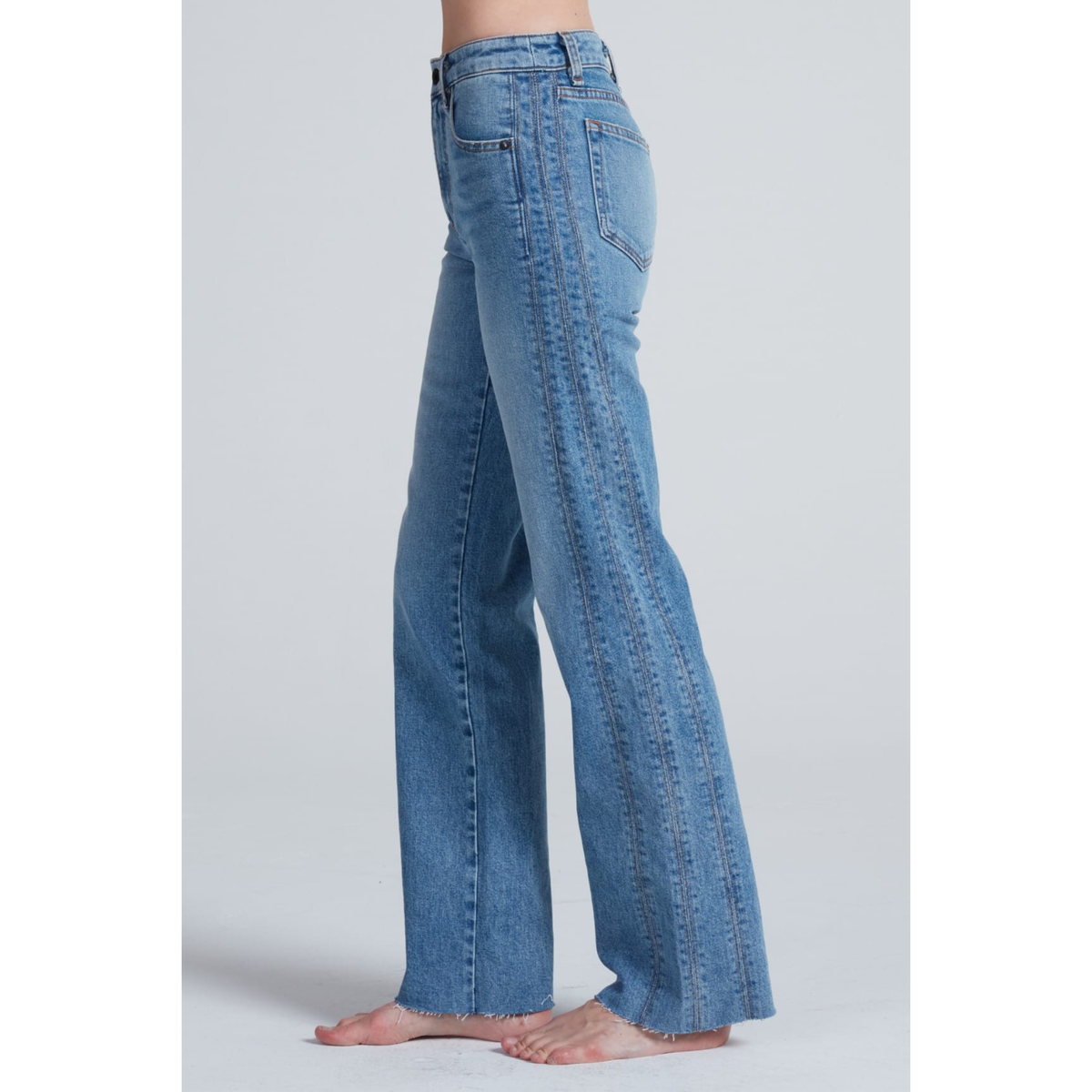 A side profile view of a person wearing high-waisted, wide-leg blue denim jeans, shown barefoot against a light gray studio background.