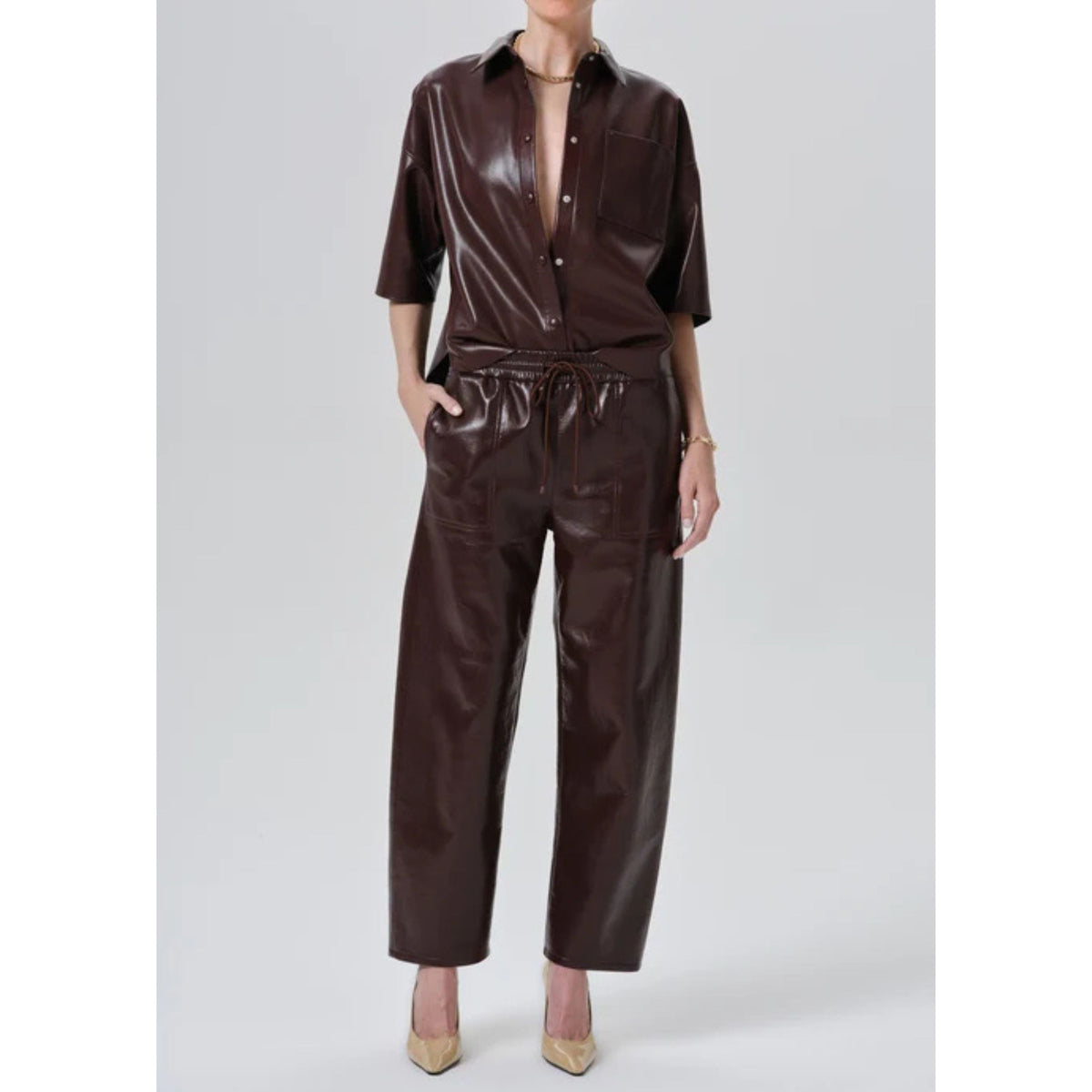 A model wearing a dark brown faux leather two-piece set consisting of a short-sleeve button-up shirt and matching wide-leg cropped pants with a drawstring waist, paired with beige pointed-toe heels against a light gray studio background.
