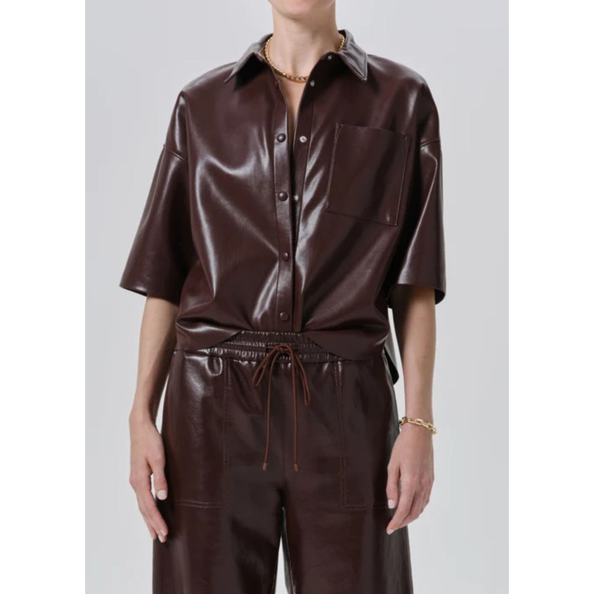 A model wearing a dark brown faux leather button-down shirt and matching drawstring trousers against a light gray studio background.