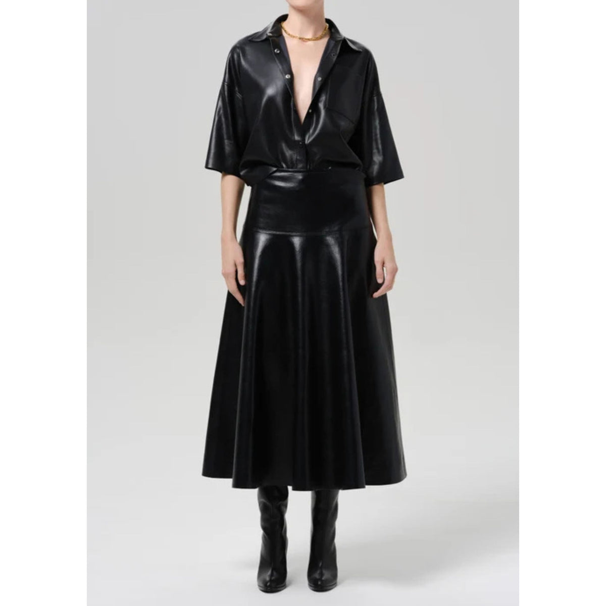 A model wearing a black faux-leather short-sleeve button-up shirt and a matching black faux-leather midi skirt, paired with black boots, against a light gray studio background.