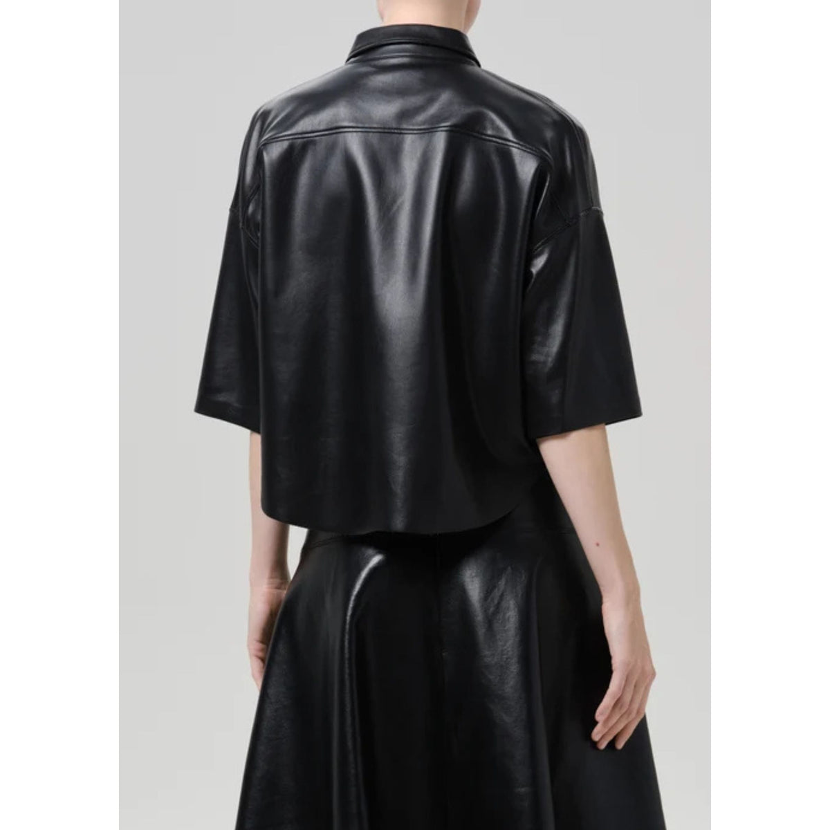 A model shown from the back wearing a black leather short-sleeve shirt and a matching black leather skirt against a light gray studio background.