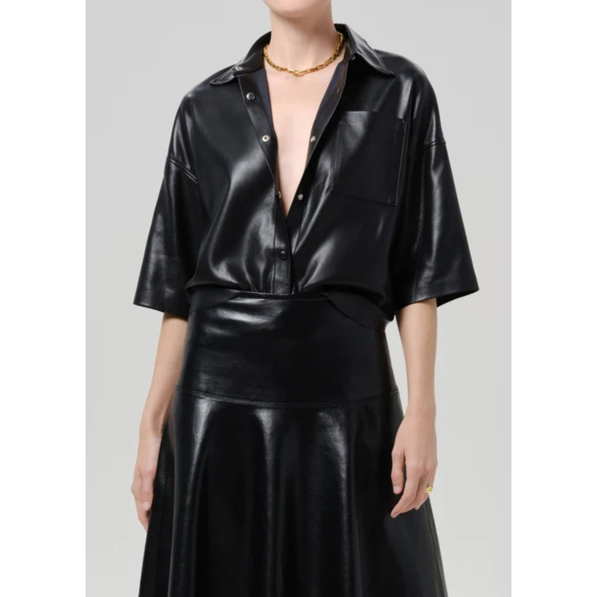 A model wearing a black faux leather short-sleeve button-up shirt and matching high-waisted skirt against a light gray studio background.