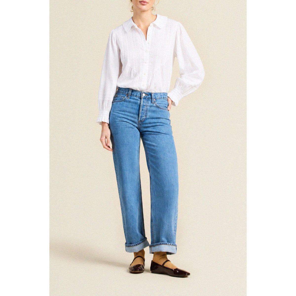 A model wearing a white long-sleeve button-down shirt and light blue wide-leg jeans with rolled cuffs, paired with dark brown flat shoes, against a solid beige background.