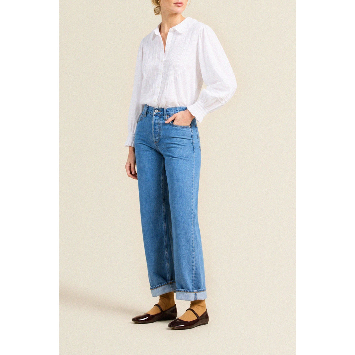 A model wearing a white long-sleeve button-up blouse and high-waisted, wide-leg blue jeans with rolled cuffs, paired with dark brown flat shoes, set against a plain beige studio background.
