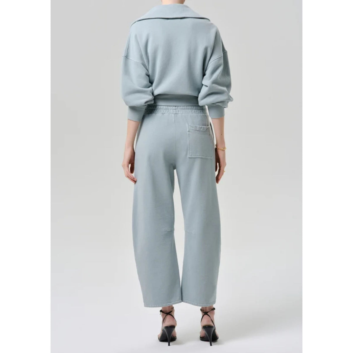 Full-length back view of a model wearing the matching light sage green sweatshirt and cropped sweatpant set paired with black heels.