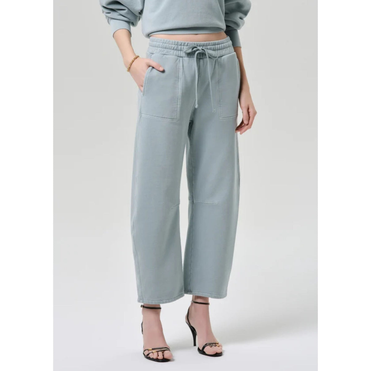Three-quarter view from the waist down showing the elastic drawstring waistband and cropped hem of the light sage green sweatpants paired with heels.