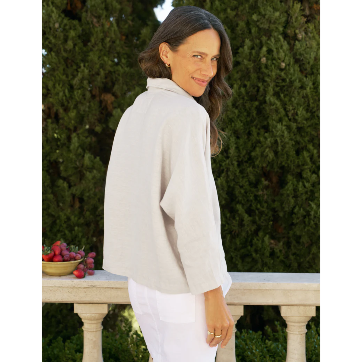 Woman wearing a light gray blouse and white pants standing outdoors with greenery in the background.