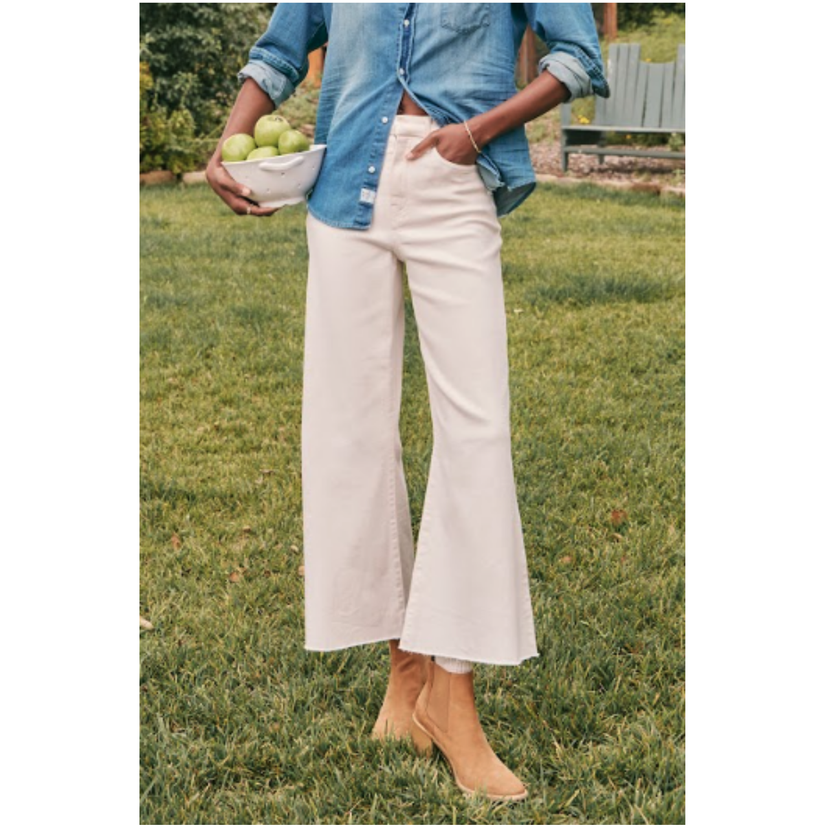A person standing on a grassy lawn wearing cream wide-leg cropped pants, a light blue denim shirt, and tan ankle boots, while holding a white bowl of green apples.