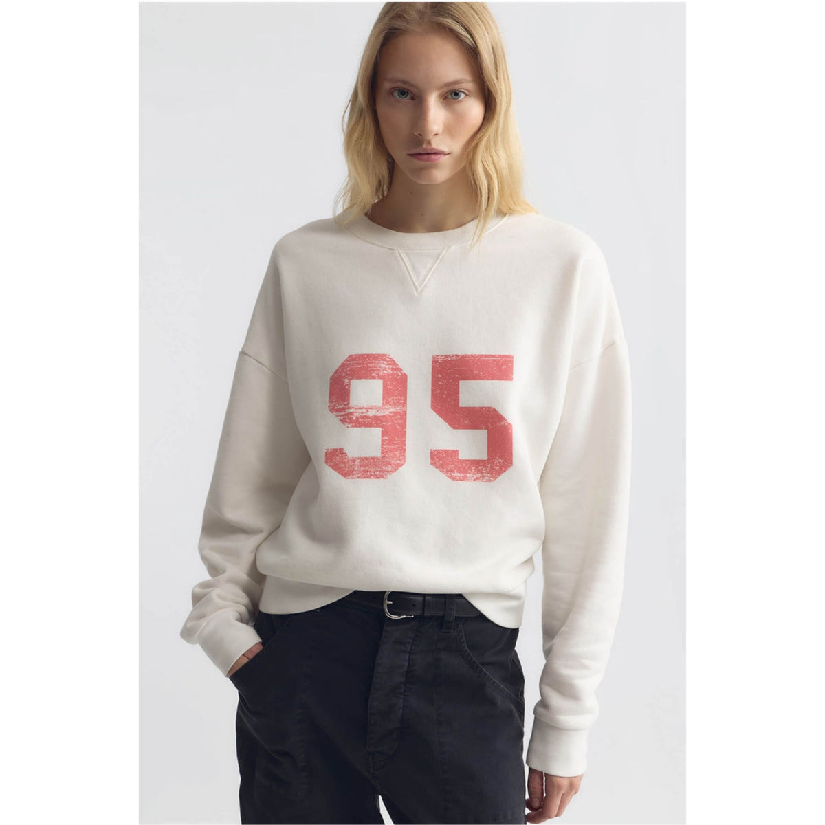 A blonde model wearing a cream-colored crewneck sweatshirt with a distressed red '95' graphic on the front, paired with dark pants, against a light gray studio background.